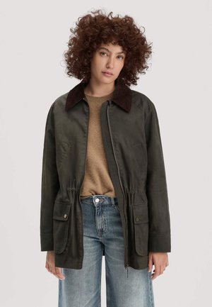 Person mit lockigem Haar, die eine dunkelgrüne Jacke mit großen Vorderentaschen, einen braunen Pullover und hellblaue Jeans trägt.