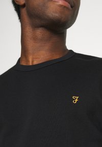 T-shirt in cotone nero con scollatura a girocollo. Presenta un piccolo logo giallo ricamato sul petto. Texture liscia, design semplice.