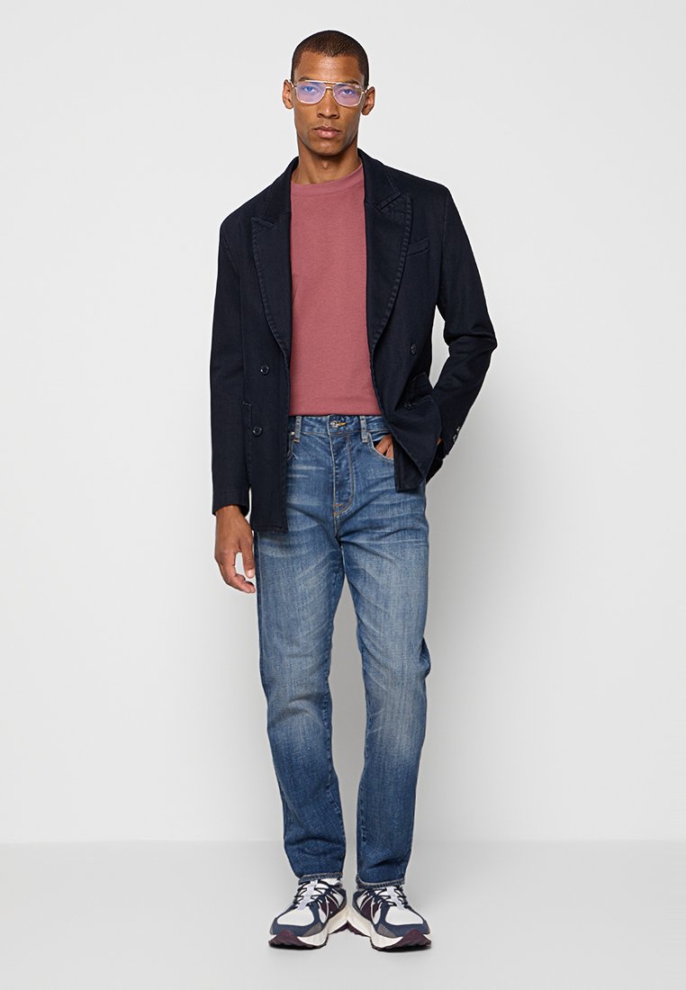 Uomo che indossa una maglietta rosa a girocollo, un blazer nero, jeans blu, sneaker e occhiali con lenti azzurre, in piedi con una mano in tasca.