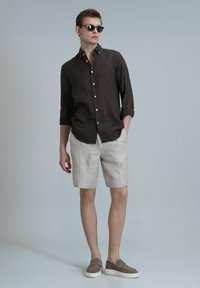 Chemise marron en lin à boutons, short en lin beige clair et chaussures plates grises. Le modèle porte des lunettes de soleil noires, dans une pose décontractée sur fond neutre.