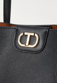 Geantă de piele neagră cu un finisaj texturat, accesorii aurii și un capac cu un închizător vizibil. Logo-ul mărcii este embosat pe capac.