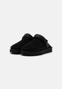 Black suede clogs with a rounded toe and a black strap across the instep. Textured surface and cushioned black rubber sole.