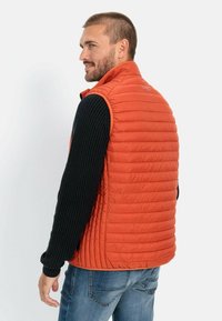 Orangen Isolierwesten mit gestepptem Muster und hohem Kragen, über einem schwarzen gerippten Langarmshirt getragen, von hinten gesehen.