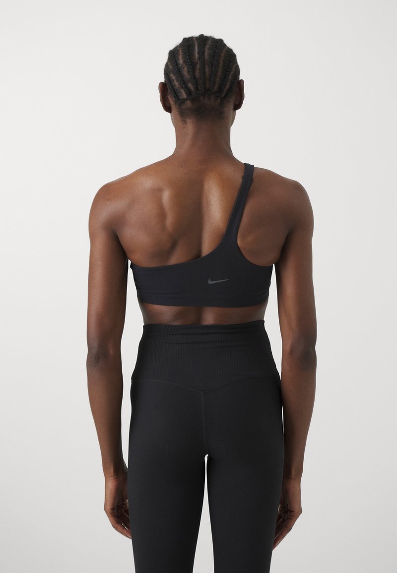 Reggiseno sportivo nero con design asimmetrico a spalla unica, caratterizzato da una superficie liscia e dettagli del logo discreti, abbinato a leggings neri a vita alta.