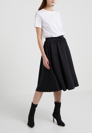 Femme portant une chemise blanche à manches courtes, une jupe noire longueur genoux avec taille à cordon, et des bottines noires à talons hauts, debout sur un sol blanc.