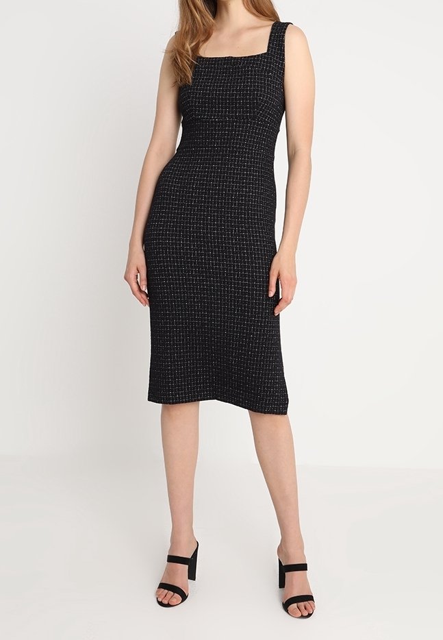 Femme portant une robe noire texturée sans manches, longueur genou, avec un décolleté carré et des sandales à talons ouvertes noires, debout devant un fond blanc.