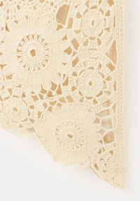 Tissu crocheté couleur crème avec des motifs floraux et des détails ajourés, montrant un coin avec des bords festonnés.