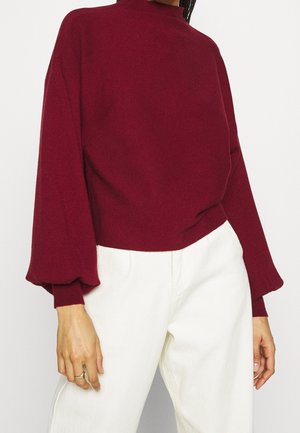 Burgundy stickad tröja med ballongsärmar, hög halsringning och ribbade ärmslut, kombinerad med vita byxor med en mjuk textur.