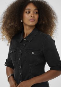 Schwarze Denim-Bluse mit sichtbaren Nähten, zwei Brusttaschen und Knopfverschluss. Verfügt über hochgekrempelte Ärmel und ein tailliertes Design.