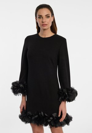 Frau trägt ein schwarzes Kleid mit langen Ärmeln und großen gerafften Blumenapplikationen an Ärmeln und Saum vor einem schlichten hellen Hintergrund.