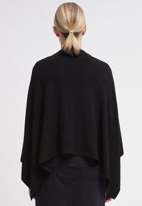 Persoon met blond haar in een lage paardenstaart, gekleed in een zwart capeachtig kledingstuk en donkere broek, staande tegen een effen lichte achtergrond.