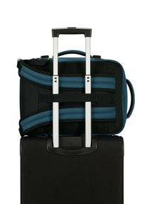 Svart resväska med en teleskophandtag, toppad med en blå och svart ryggsäck som är fäst med band, och har en vadderad rygg och nätdetaljer.