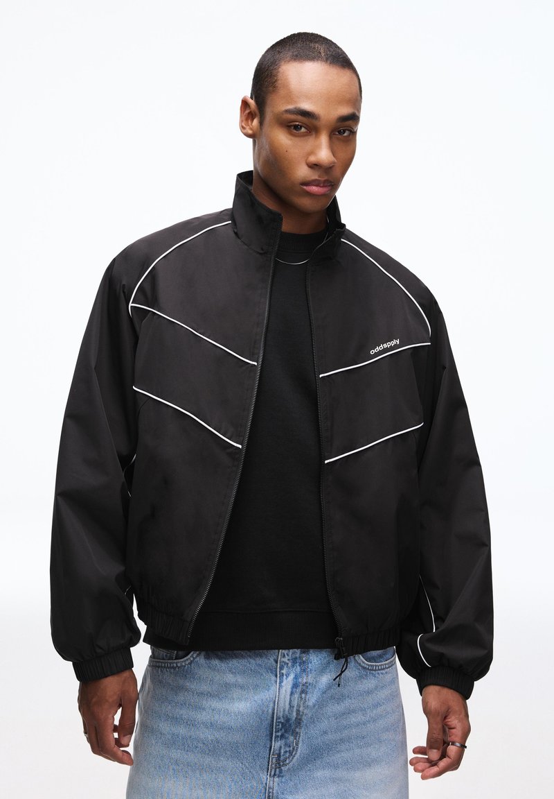 Black zip-up jacket with white accent lines. Features a high collar, elastic cuffs, and a relaxed fit. Worn over a black sweater.