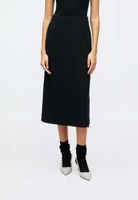 Svart midi-skjørt med et glatt, elastisk stoff; har en tettsittende midje og går til midt påleggen. Kombinert med lyse spisse hæler og sorte sokker.