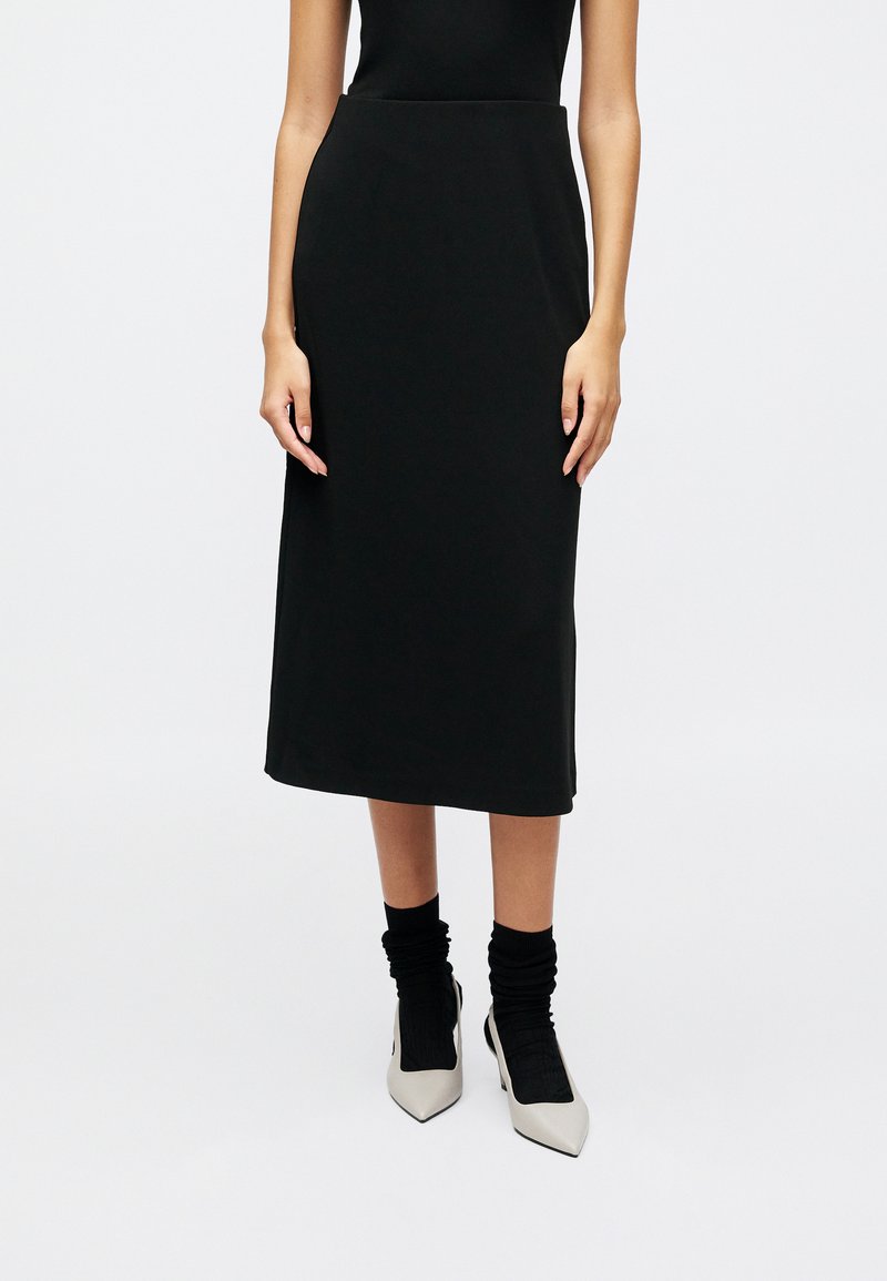 Svart midi-skjørt med et glatt, elastisk stoff; har en tettsittende midje og går til midt påleggen. Kombinert med lyse spisse hæler og sorte sokker.