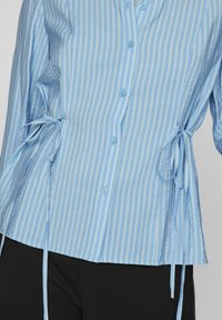 Camisa de rayas azul claro con mangas largas, que presenta cierre de botones, lazos en los laterales y un ajuste relajado. Tejido suave con un brillo sutil.