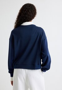 Sudadera de color azul marino con cuello blanco, de corte holgado y puños de canalé. Presenta una textura suave y un dobladillo corto en la parte posterior.