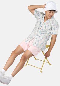 Chemise à manches courtes avec un léger motif floral dans des couleurs pastel, associée à un short rose et des baskets blanches, assise sur une chaise pliante jaune.