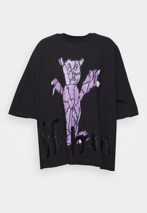 Camiseta negra oversize con un gran dibujo abstracto de oso morado y texto negro parcialmente visible a lo largo del dobladillo inferior.