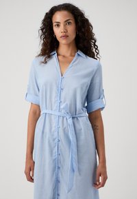 Vestido riscado azul claro com mangas curtas, frente abotoada e cinto amarrado na cintura. Apresenta um colarinho e punhos de mangas enrolados.