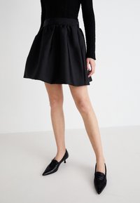 Saia preta evasé com uma textura suave e cintura franzida, combinada com sapatos de salto agudo pretos e uma blusa preta de mangas longas.