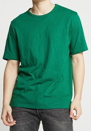 Homme portant un T-shirt vert uni à manches courtes et un jean gris foncé, debout devant un fond clair.