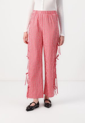 Rood-witte geruite broek met wijde pijpen en strikjes aan de zijkant, gedragen met zwarte platte schoenen en een wit langmouwig hemd met knopen.