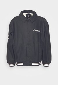Veste noire Converse à boutons avec logo blanc brodé sur la poitrine et poignets et taille rayés, présentée sur un fond uni.