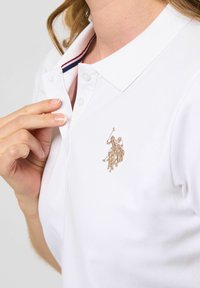 Vit pikétröja med krage, som har en placket med tre knappar och en beige broderad logotyp på vänster sida. Inklusive röd och blå randig detaljering.