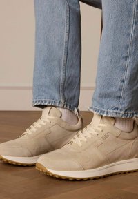 Beige Wildleder-Sneaker mit weißen Sohlen, strukturiertem Obermaterial und flachen Schnürsenkeln, kombiniert mit hellblauen, verkürzten Jeans, die weiße Socken zeigen.