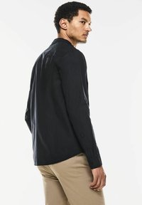 Schwarzes Langarmshirt mit Kragen, taillierter Passform und Knopfverschluss an den Manschetten, von hinten vor einem weißen Hintergrund gezeigt.