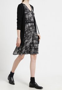 Cardigan nero sopra un abito grigio a fantasia con scollatura a V e gonna svasata, abbinato a scarpe nere e calzini scuri.