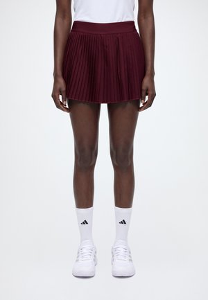 Modèle portant une jupe plissée bordeaux, un haut blanc, des chaussettes montantes et des baskets blanches sur un fond de studio uni.