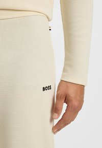 Haut thermique beige avec une surface texturée et côtelée. Présente un logo noir "BOSS" sur le côté gauche. Coupe slim, manches longues.