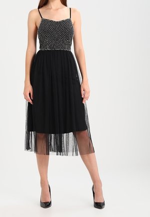 Kobieta w czarnej sukience midi z koralikowym gorsetem, cienkimi ramiączkami i przezroczystą tiulową nakładką, w czarnych szpilkach.