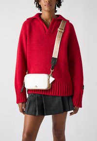 Maglione rosso a maglia con colletto, abbinato a una gonna a quadri. Una piccola borsa a tracolla bianca con dettagli dorati e una forma triangolare pende dalla spalla.