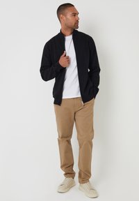 Giacca nera con zip sopra una t-shirt bianca, abbinata a pantaloni khaki e sneakers beige. Tessuto liscio, design slim, in posizione eretta.