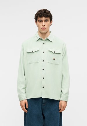 Νέος άνδρας με κοντά σγουρά μαλλιά, φορώντας ένα ανοιχτό πράσινο πουκάμισο από κορδόνι με κουμπιά και φαρδιά σκούρα μπλε τζιν, στέκεται κοιτάζοντας μπροστά.