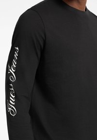 Sort langærmet t-shirt lavet af bomuld med rund hals. Kan prale af "Guess Jeans" i hvid skrift langs venstre ærme.