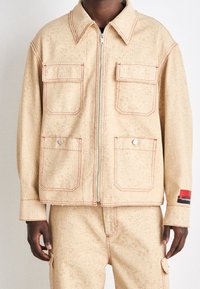 Beige jacka med ton-i-ton-mönster, två bröstfickor, röd sömnad och en frontdragkedja. Vänster ärm har en svart och röd märkesetikett.