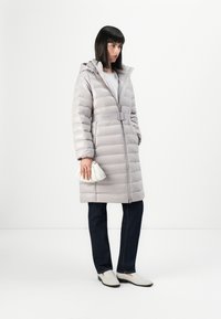Lys grå pufferjakke med hette, belte i midjen og horisontalt quiltet tekstur. Kombinert med mørke jeans og hvite slip-on sko.