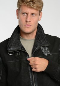 Sorte læderjakke med pelskrave, lynlås fortil og dekorativt hardware. Har en tekstureret overflade og tætsiddende manchetter.