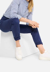 Kobieta siedzi na białym bloku, ma na sobie niebieską koszulę w paski, granatowe spodnie podwinięte na kostkach i białe trampki, z dłońmi złożonymi.