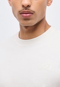 Κοντινό πλάνο κάτω μέρους προσώπου και λαιμού άνδρα που φοράει λευκό μπλουζάκι EA7 Emporio Armani με διακριτικό λογότυπο στο στήθος.