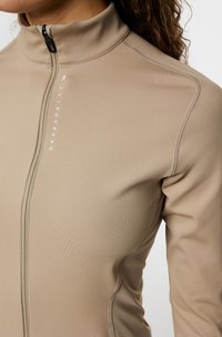 Beige zip-up jacka med hög krage, slät textur, kontrasterande sömmar och vit logodetalj längs dragkedjan. Åtsittande design.