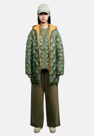 Modèle portant un manteau matelassé vert sauge avec une doublure orange, une casquette assortie, un haut olive et un pantalon à jambes larges, ainsi que des chaussures beiges.