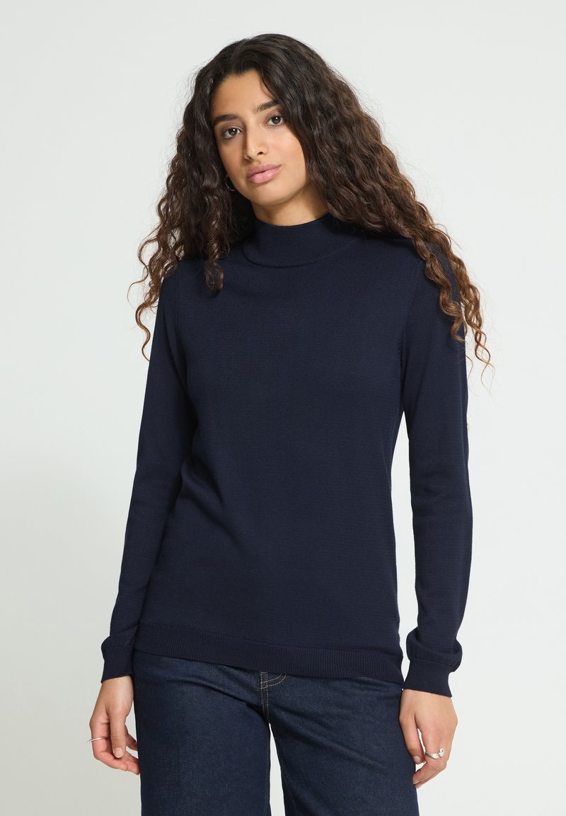 Femme aux longs cheveux bouclés portant un pull à col roulé bleu marine et un jean bleu foncé, debout devant un fond clair uni.