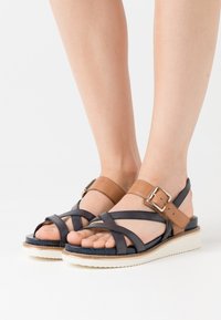 Braune und marineblaue Leder-Sandalen mit überkreuzten Riemen, verstellbarem Knöchelverschluss und weißer Gummisohle. Strukturiertes Obermaterial mit offener Zehenpartie.