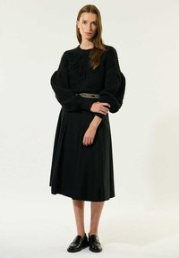 Črn, teksturiran pulover z obsežnimi rokavi, v kombinaciji s tekočo črno krilom. Model nosi črne loaferje in pas z napisanim okrasjem.