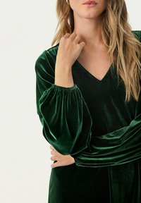 Smaragdgrünes Samtkleid mit V-Ausschnitt, puffärmeligen Ärmeln und tailliertem Oberteil, das eine weiche Textur zeigt.